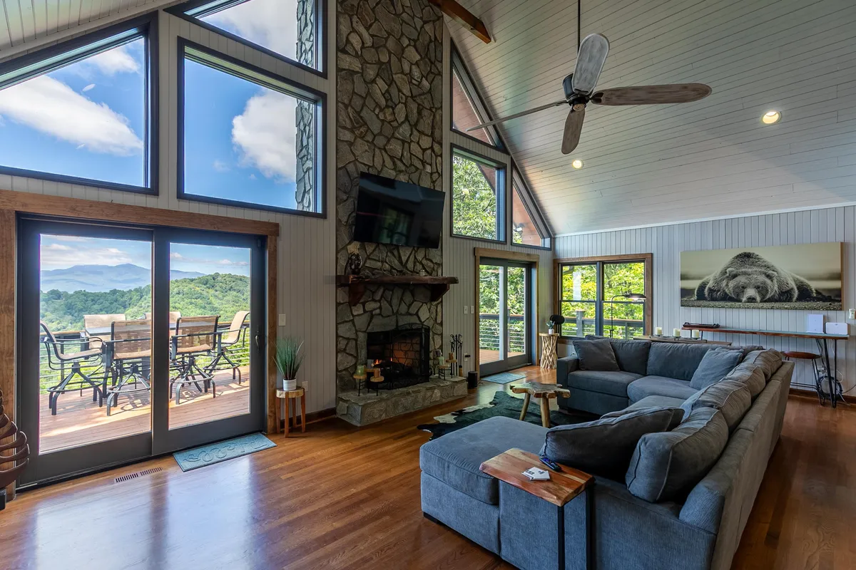 A spacious living room featuring a dramatic 20-foot stone fireplace, a large sectional sofa, and expansive cathedral-height windows that provide sweeping mountain views. The deck is visible through the windows, further connecting the interior with the scenic outdoors.