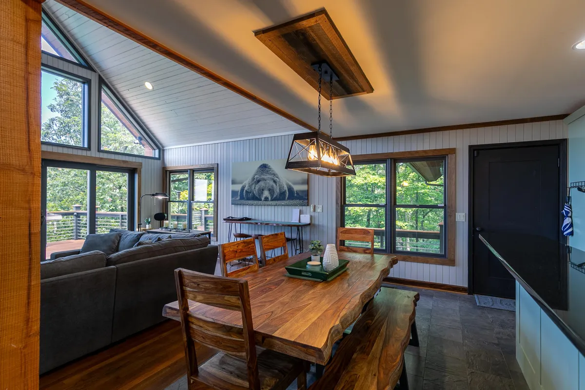 A bright interior dining area with large windows framing views of the outdoors. The room includes a dining table, comfortable seating, and inviting décor. Sunlight streams in, creating a warm atmosphere ideal for working remotely or enjoying a relaxing meal.