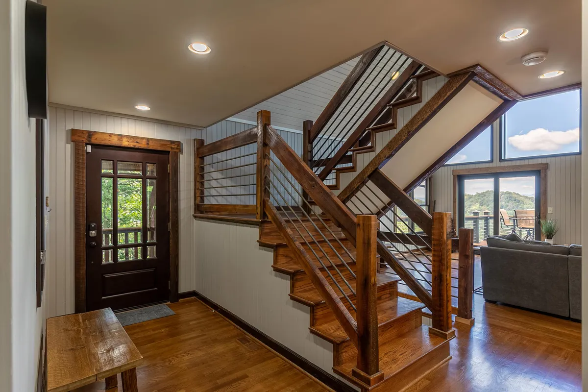 The image captures the entryway staircase, front door, and the flood of natural light from windows that reveal glimpses of the outdoors. There’s a subtle sense of transition from indoors to nature.