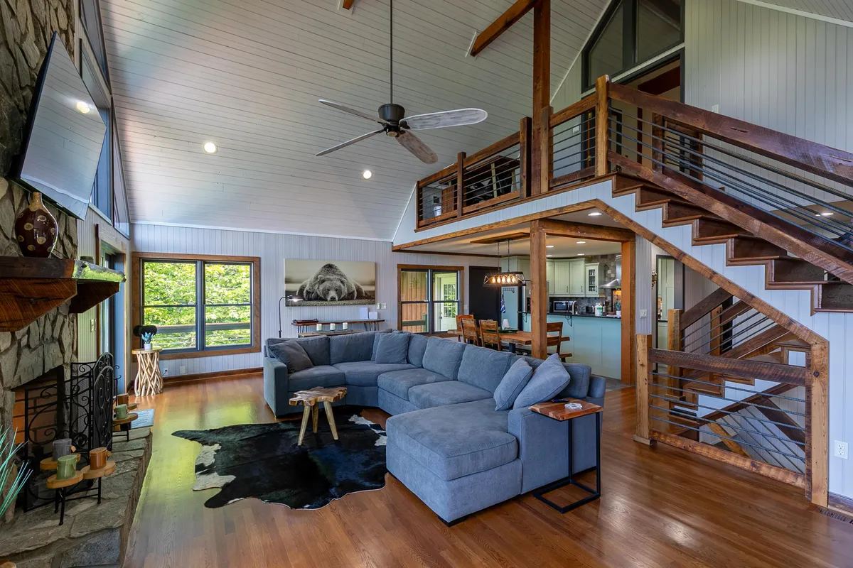 This image features a spacious living area with a large sectional sofa, a stone fireplace, and expansive windows that fill the room with natural light. The windows provide views of the surrounding forest, hinting at the outdoor spaces accessible from the home.
