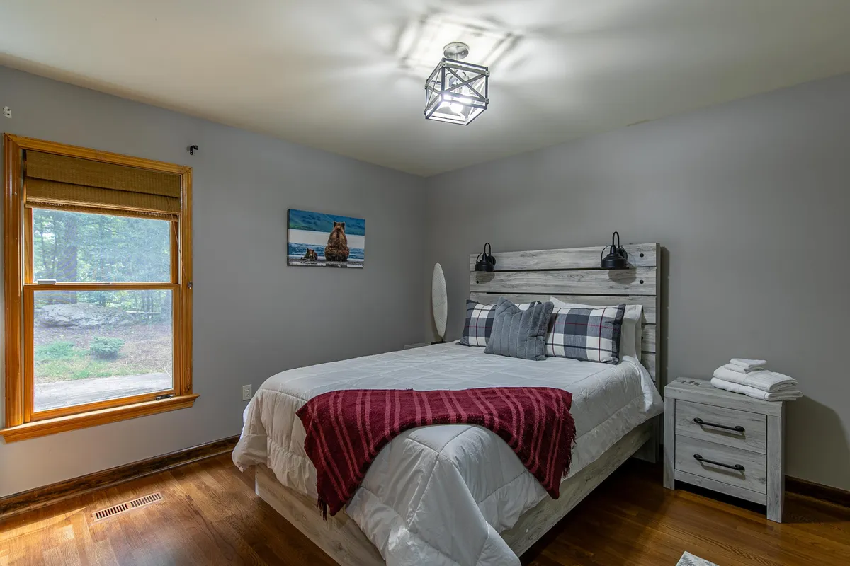 A cozy queen bedroom with plaid bedding, wooden bedside table, framed artwork, and a window letting in natural light. The space exudes calm and comfort, ideal for restful sleep or quiet reading.