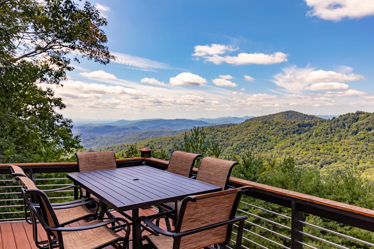 A spacious wooden deck featuring a dining table and chairs, set against a stunning backdrop of rolling mountains and expansive sky. Lush greenery surrounds the deck, creating an inviting space for outdoor gatherings and meals.