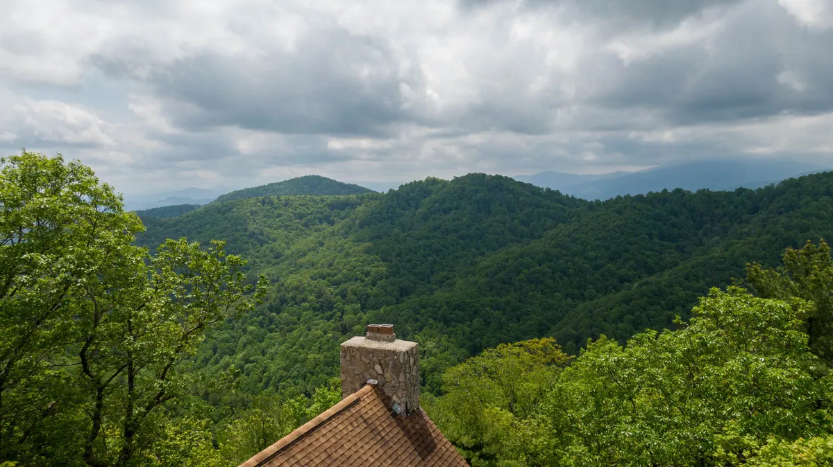 A wide view of a mountain range covered in lush greenery, with the house's chimney just visible in the foreground. The scene evokes a strong sense of place, with expansive vistas perfect for hiking, wildlife spotting, and enjoying nature's tranquility.