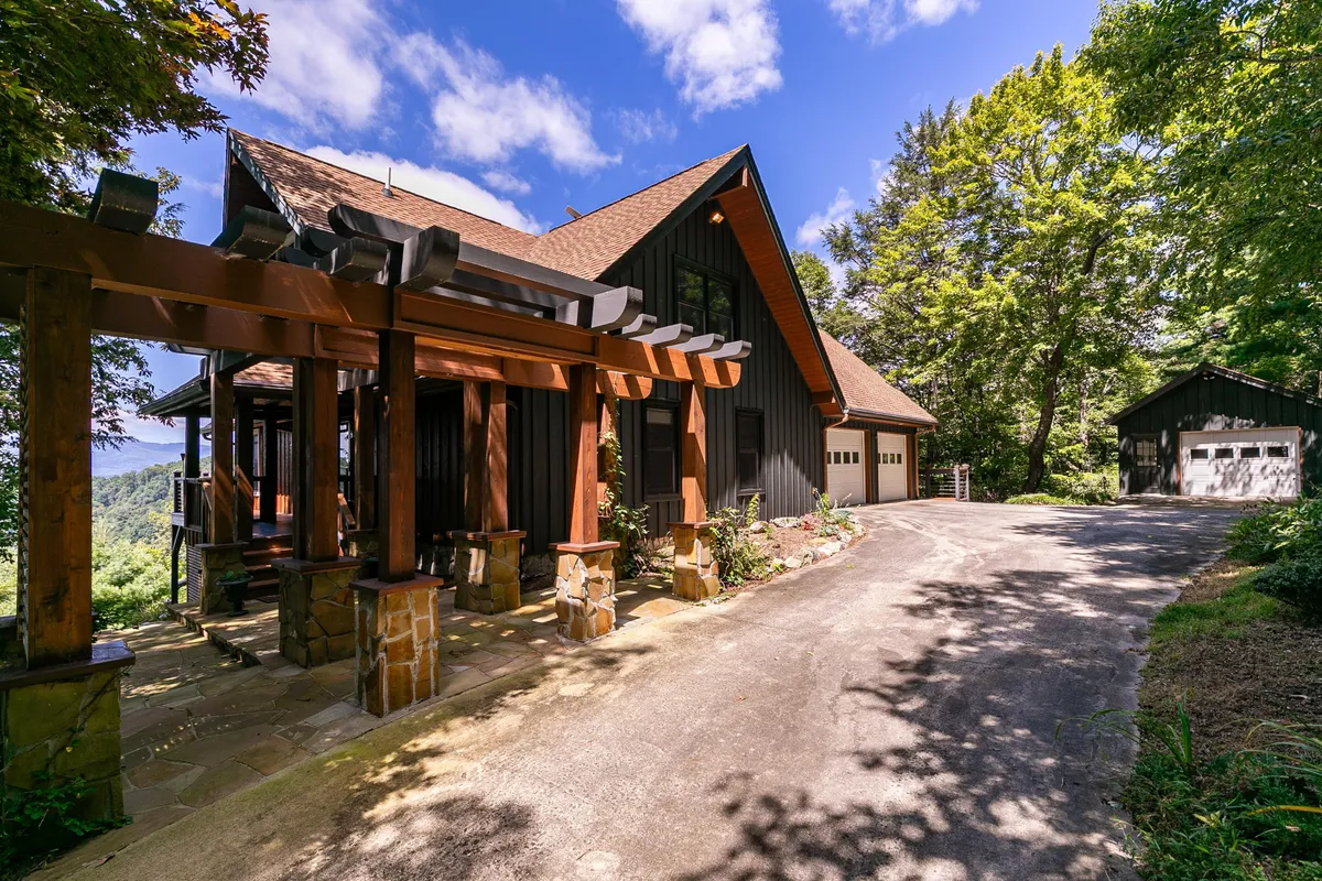 The image shows the exterior of a modern mountain home framed by a wooden pergola and surrounded by lush greenery. A stone pathway leads through the landscaped yard, inviting guests toward the home. The scene exudes tranquility, with natural forest and shrubbery blending seamlessly into the architecture. The view hints at expansive mountain vistas, perfect for relaxing outdoors or gathering with loved ones.