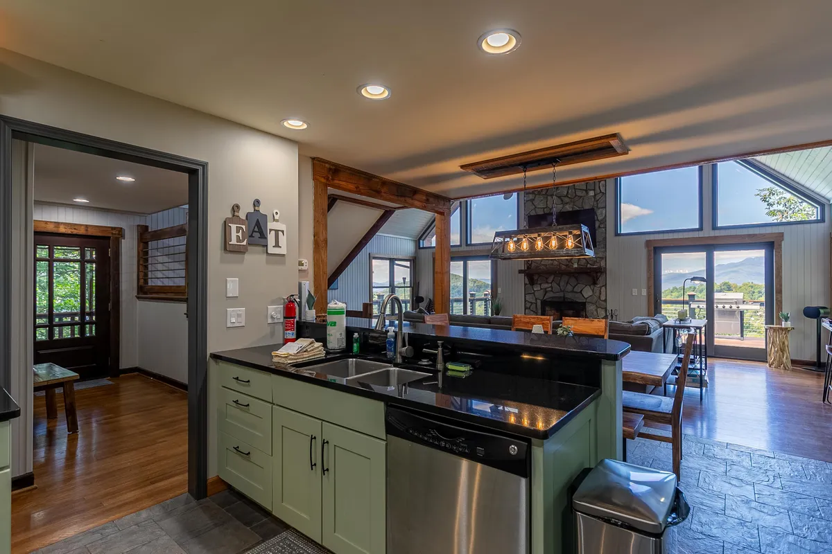 An open-concept kitchen, dining, and living area with a prominent kitchen island, solid wood dining table, granite countertops, and a view of the stone fireplace and large windows. The space exudes a communal and welcoming atmosphere, ideal for gatherings and shared meals.