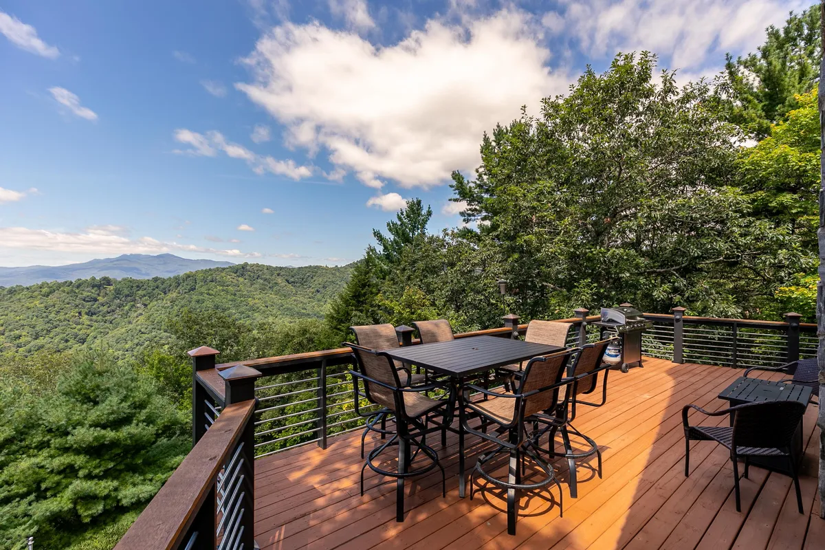 An outdoor dining area set on a spacious deck overlooks majestic mountain views and is surrounded by lush green trees. The tranquil setting invites guests to dine al fresco, relax with morning coffee, or stargaze in the evening, with ample space for gatherings.