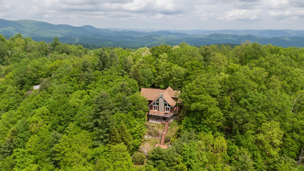 A peaceful exterior view of the house nestled among tall trees, with the mountain range soaring in the background. The scene captures the home's harmonious integration with its natural surroundings and the grandeur of the mountain landscape.