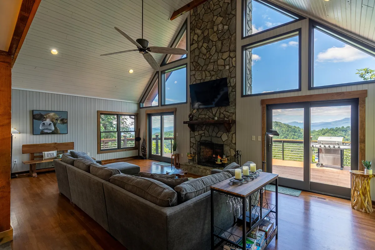 This image showcases another angle of the living area, emphasizing the large stone fireplace, comfortable sectional sofa, and expansive windows looking out onto the forested landscape.