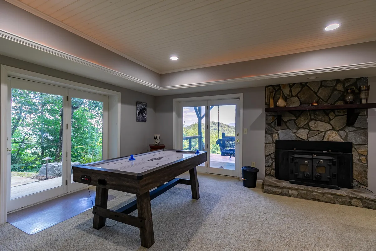 A playful, cozy recreation room featuring an air hockey table, a stone fireplace, and large windows that offer views of the outdoors. The space is furnished for games and relaxation, blending fun and comfort.