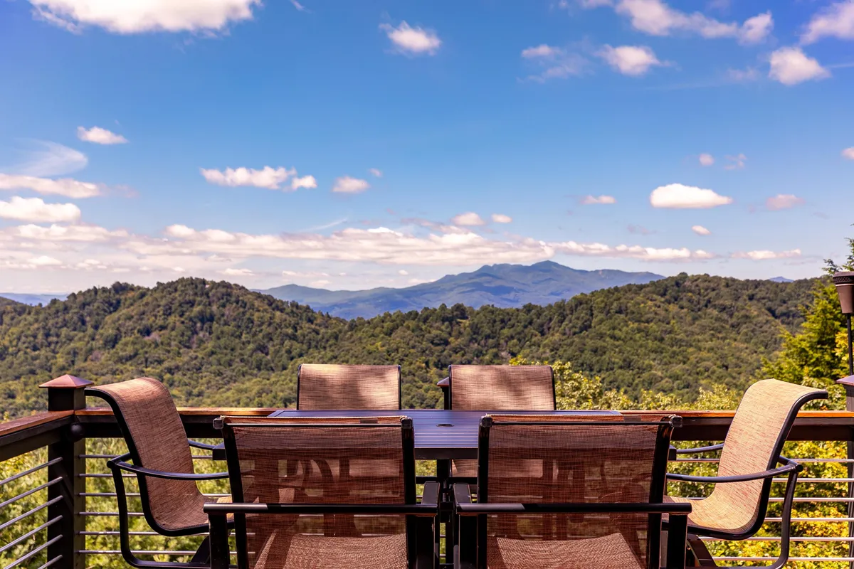 An outdoor dining table and chairs are set on a deck or patio, overlooking expansive mountain views. The scene is tranquil, with the table invitingly placed to take advantage of the vista. The natural setting suggests an inspiring space for meals, work, or relaxation, with the mountains providing a dramatic and peaceful backdrop.